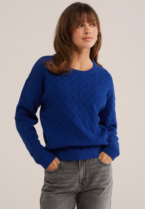 Mujer con cabello castaño hasta los hombros, vistiendo un suéter azul real texturizado y vaqueros grises con las manos en los bolsillos, mirando de lado.