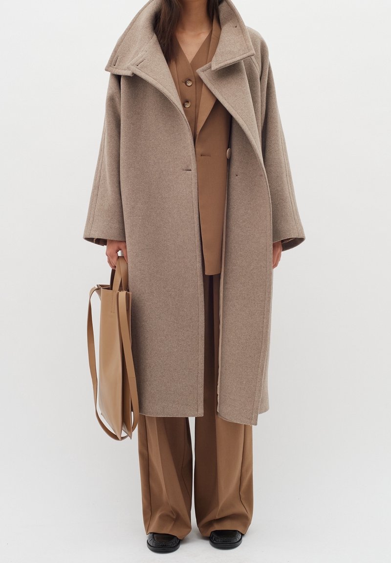 Cappotto di lana beige con maniche ampie, colletto asimmetrico e due bottoni frontali; abbinato a un completo marrone e a una borsa a tracolla in pelle beige.