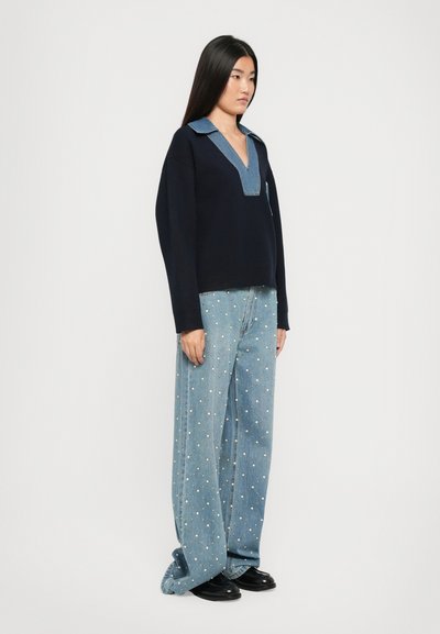 Maglione blu marino con colletto in denim, abbinato a jeans a gamba larga di un blu chiaro con decorazioni in perline bianche. Scarpe nere.