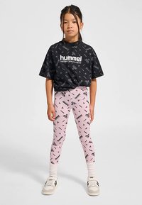 Svart t-shirt med "hummel PERFORMANCE"-logotyp och upprepande mönster. Rosa leggings med svarta motiv. Modellen bär vita sneakers och strumpor.
