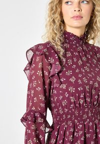 Burgundowa sukienka w kwiaty z falbaniastego rękawa, gumką w talii i wysokim dekoltem. Posiada małe białe i zielone wzory kwiatowe na przezroczystym materiale.