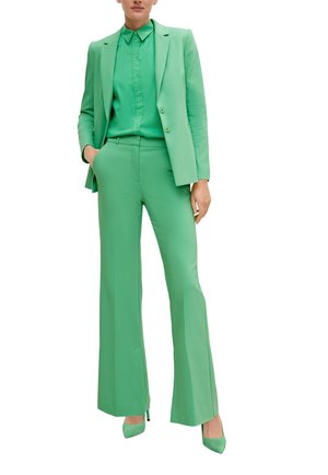 Completo in poliestere verde composto da un blazer aderente e pantaloni svasati, con una camicia button-up sotto. Tacchi alti verdi abbinati.