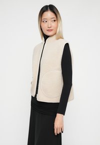 Mladá žena so rovnými blond vlasmi, oblečená v bezrukávovej béžovej fleecovej vestě nad čiernym dlhým rukávom a sukňou, pozerajúca sa do strany.
