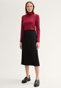 Rotes Rollkragenoberteil aus dehnbarem Material, kombiniert mit einem schwarzen plissierten Midirock und klobigen schwarzen Slippern mit glänzendem Finish.