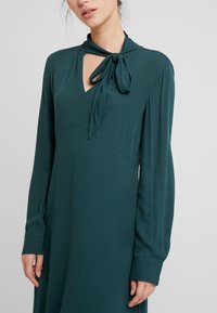 Vestido de manga larga en azul verdoso profundo con escote en forma de llave, detalle de cuello atado y tela suave. El diseño presenta un cuerpo ajustado y una falda fluida.