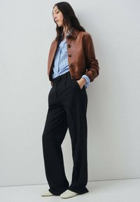 Veste bomber en cuir marron avec de gros boutons, portée sur une chemise rayée bleue et blanche et un pantalon large à rayures. Le tout est assorti de chaussures blanches.