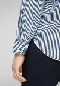Camicia a maniche lunghe a righe blu e bianche con dettagli sui polsini abbottonati, abbinata a jeans scuri. Tessuto di cotone morbido, vestibilità sagomata.
