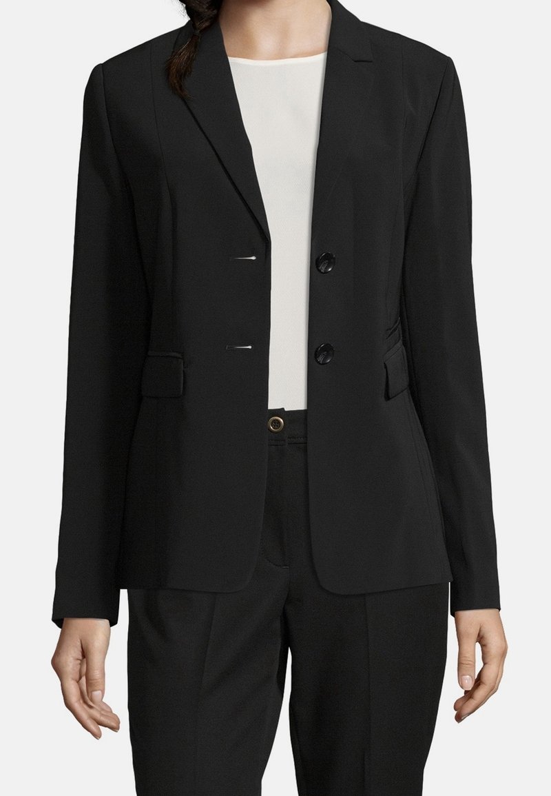 Femme portant un blazer noir ajusté sur un haut blanc et un pantalon noir devant un fond uni.