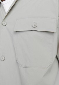 Helles graues Hemd mit Knopfleiste und einer einzigen Brusttasche, ausgestattet mit glatter Textur und schlichter Optik, geschlossen mit passenden Knöpfen.