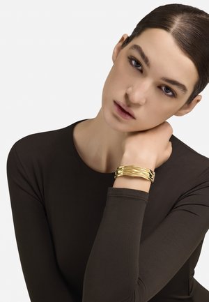 Arany csavart karkötő egy csuklón, sima fémes textúrával. A háttér fehér, a viselt ruha sötétbarna.