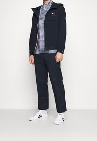 Hombre vestido con una chaqueta azul marino, una camisa de cuadros azul y blanca, pantalones azul marino y zapatillas blancas con logo de estrella negra, de pie frente a un fondo blanco.