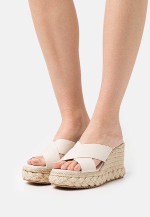 Fødder iført beige platform wedge sandaler med krydssnoede stropper og flettede reb-texture såler på en hvid baggrund.