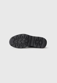 Semelle de chaussure en caoutchouc noir avec un motif texturé, comportant des rainures profondes et une section centrale lisse. Avant arrondi et bords plats.