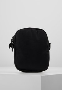 Schwarze Stofftasche in quadratischer Form, mit einem Reißverschluss und seitlichen Hardware-Akzenten. Die Textur ist glatt und matt.