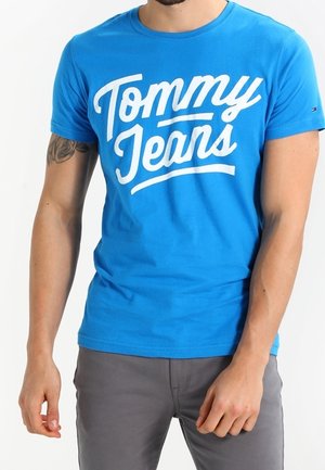 Mężczyzna w jasnoniebieskiej koszulce z krótkim rękawem z dużym białym napisem „Tommy Jeans” oraz w szarych spodniach na białym tle.