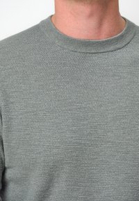 Ljust grå tröja med ribbad rund halsringning, med ett texturerat stickmönster. Materialet ser mjukt och medelviktigt ut.