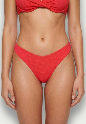 Tors kobiety ubrany w fakturowany czerwony dół od bikini i pasujący top, stojąca z rozluźnionymi ramionami wzdłuż ciała na neutralnym tle.