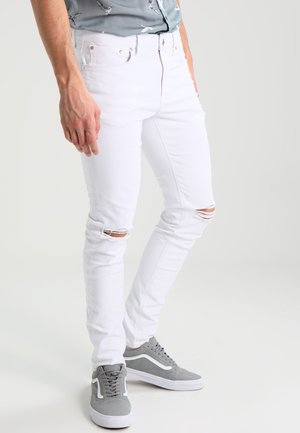 Uomo che indossa jeans bianchi aderenti con strappi sulle ginocchia e sneakers grigie, in piedi contro uno sfondo chiaro e uniforme.