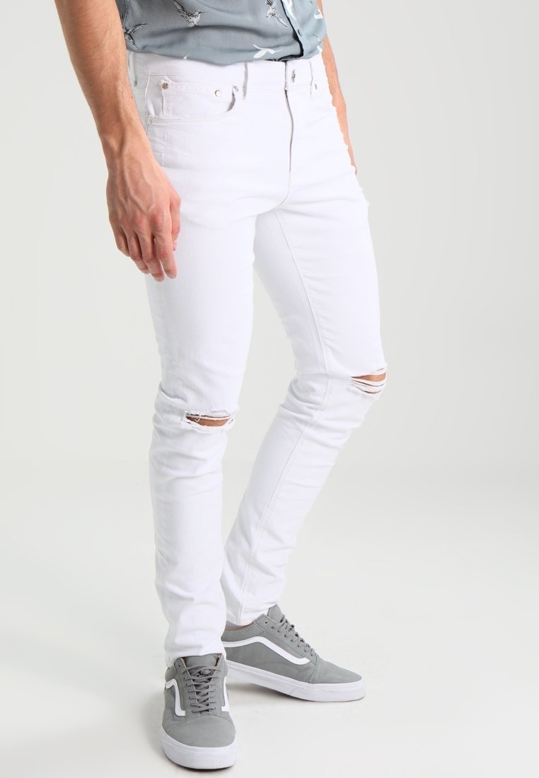 Uomo che indossa jeans bianchi aderenti con strappi sulle ginocchia e sneakers grigie, in piedi contro uno sfondo chiaro e uniforme.