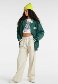 Giacca bomber verde con polsini a righe, top corto bianco con stampa grafica, pantaloni larghi beige e un berretto verde brillante.