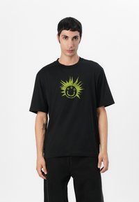 Black cotton t-shirt featuring a green sun design with a smiley face and the letters "HGO" above, short sleeves, and a relaxed fit.