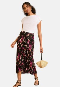 Melna maxi svārku ar ziedu rakstu rozā un krēmkrāsā, apvienota ar baltu T-kreklu, un papildināta ar melnām sandales ar siksniņām un austu ventilatora somu.