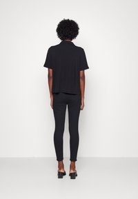 Personne aux cheveux courts et bouclés portant une chemise noire à manches courtes, un pantalon noir ajusté et des sandales à talons noires, debout de dos.