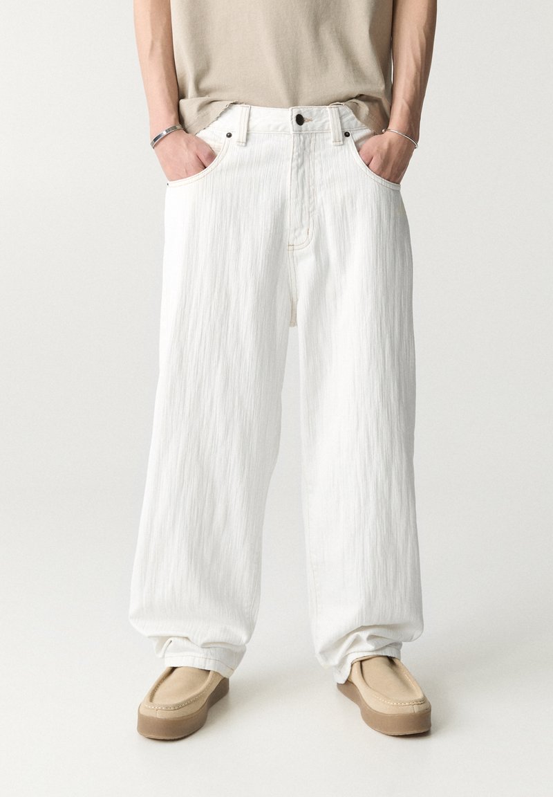 Jean blanc à jambes larges avec une finition texturée, un design à cinq poches, une fermeture à bouton et des détails de couture subtils, associés à des chaussures beiges à enfiler.