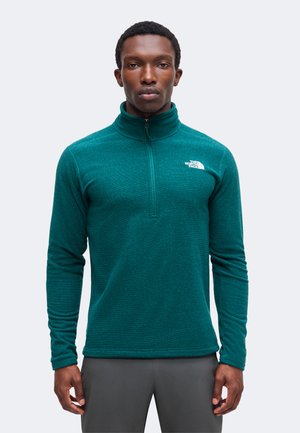 Homme portant un haut polaire à manches longues bleu sarcelle avec fermeture éclair quart de zip et logo The North Face sur la poitrine, debout devant un fond uni.