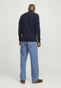 Marineblauer Rippstrickpullover mit hohen Kragen, kombiniert mit hellblauen, locker sitzenden Jeans und braunen Schuhen, von hinten gesehen.