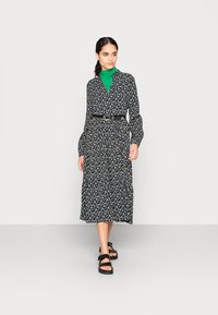 Vestido preto floral com mangas longas e decote em V, ajustado na cintura com um cinto preto. Usado sobre uma blusa de gola alta verde, combinado com sandálias pretas.