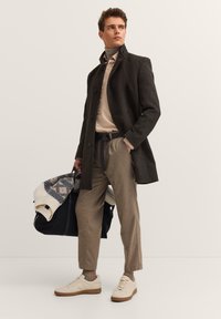 Ciemnoszary wełniany płaszcz, beżowy golf, brązowe spodnie w kratę, białe sneakersy, trzymając granatową torbę z wzorzystym swetrem narzuconym na nią.