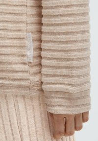 Beiges strukturiertes Gewebe mit horizontalen Streifen, Nahaufnahme eines Kleidungsstück-Ärmels. Das Etikett ist an der Naht sichtbar.