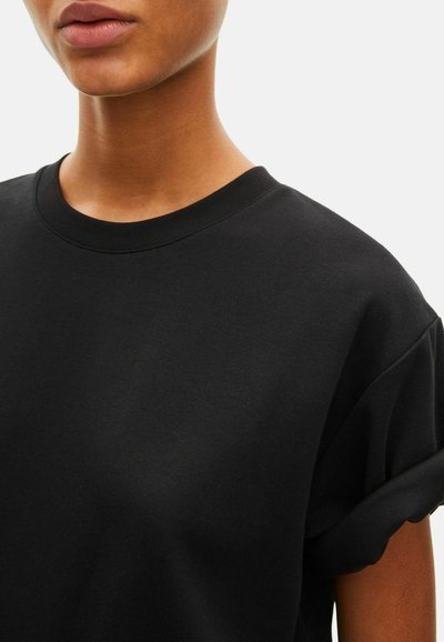 Zwart katoenen T-shirt met een ronde hals en opgerolde mouwen, met een gladde textuur en een losse pasvorm. Close-up van de torso.