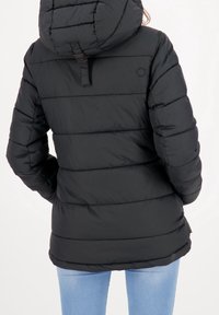 Persoon in een zwart gewatteerd jack met capuchon en horizontale stiksels, en een lichtblauwe spijkerbroek, staand met de rug naar de camera gekeerd.