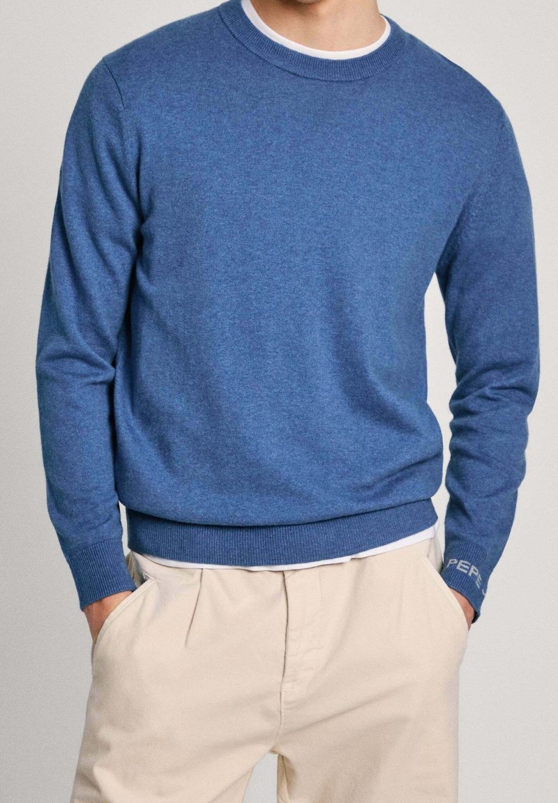 Man som bär en blå tröja med rund halsringning och beige byxor med händerna i fickorna, mot en enfärgad neutral bakgrund.