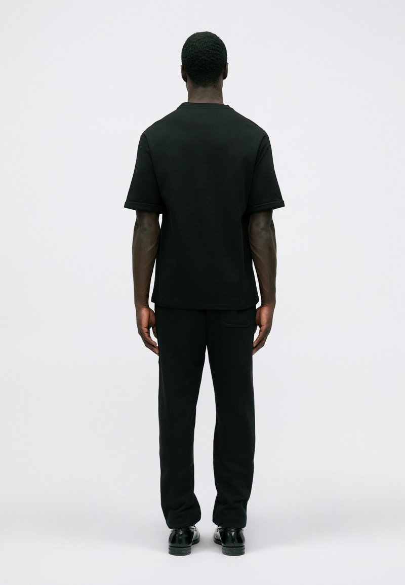 Homme se tenant de dos, portant une chemise à manches courtes noire unie, un pantalon noir et des chaussures habillées noires sur un fond blanc uni.