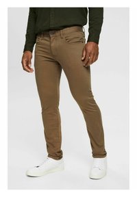 Bruine slim-fit jeans met een gladde textuur, hebben een knoopsluiting, vijf zakken en zijn gecombineerd met witte sneakers.