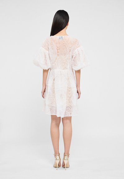Robe blanche transparente avec texture florale, manches bouffantes, taille cintrée et coupe mi-longue. Portée avec des sandales à talons dorés.