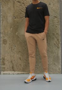 Svart Nike t-shirt med orange logotyp, beige smalpassande sweatpants och vita sneakers med orange detaljer. Avslappnad sportklädsel, minimalistisk design.