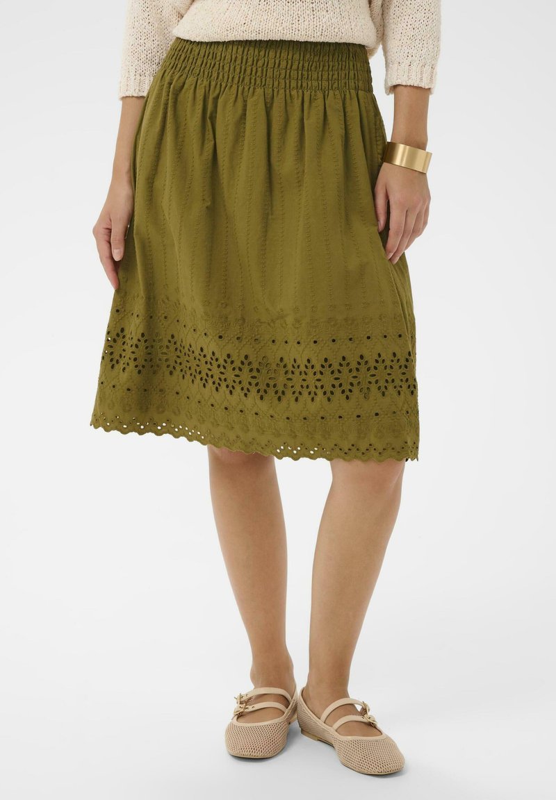 Femeie purtând o fustă verde măsliniu până la genunchi cu broderie ochiuri, pantofi bej din plasă, pulover bej tricotat și brățară aurie tip manșetă.