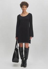 Schwarzes Strickkleid mit Rundhalsausschnitt, Glockenärmeln und Kontrastnähten am Saum, kombiniert mit kniehohen schwarzen Stiefeln und einer Tragetasche.