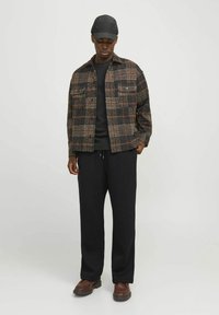 Jachetă din flanel în carouri de culoare neagră, roșie și bej, asociată cu un tricou negru și pantaloni de trening negri. Pantofi din piele maro cu talpă groasă.