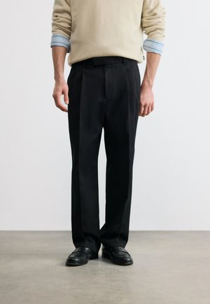 Homme portant un pantalon de costume noir, des mocassins noirs, un pull beige par-dessus une chemise bleue aux manches retroussées, debout sur un sol gris contre un mur blanc.