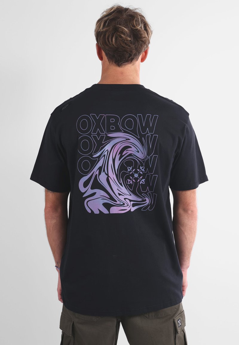 T-shirt in cotone nero con una grande grafica a onde viola e bianche e un design di testo ripetuto sul retro. Maniche corte e scollatura rotonda.