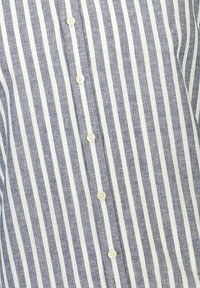 Blauw-wit gestreepte shirt met een knoopsluiting aan de voorkant en contrasterende knopen, gemaakt van textuurstof met smalle verticale strepen.