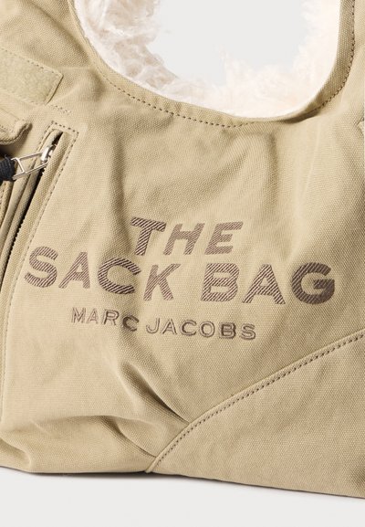 Sac en tissu beige avec "THE SACK BAG MARC JACOBS" brodé sur le devant, fermeture éclair visible et doublure blanche et duveteuse à l’intérieur.