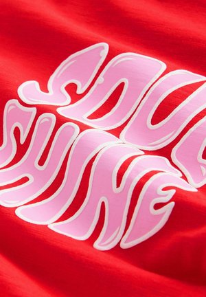 Roter Stoff mit großen, wellenförmigen, pinken und weißen umrissenen Buchstaben, die ein teilweise sichtbares Wort oder eine Phrase bilden.