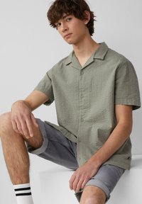Korte mouwen, knoopsluiting shirt in gestructureerde olijfgroene stof, met een notch-kragen en voorste knopen, gedragen met grijze shorts en witte sokken.