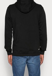 Sweat à capuche noir avec cordon de serrage, manches longues et poignets côtelés. L'ourlet inférieur est orné d'un petit logo rouge. Associé à un jean bleu.